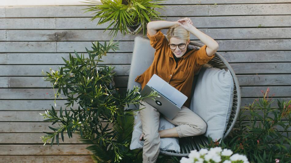 Eine Frau sitzt mit einem Laptop auf dem Schoß auf einer Veranda.