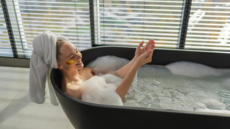 Eine Frau liegt in einer Badewanne. Sie hat goldene Pads unter den Augen und trägt einen Haarturban.