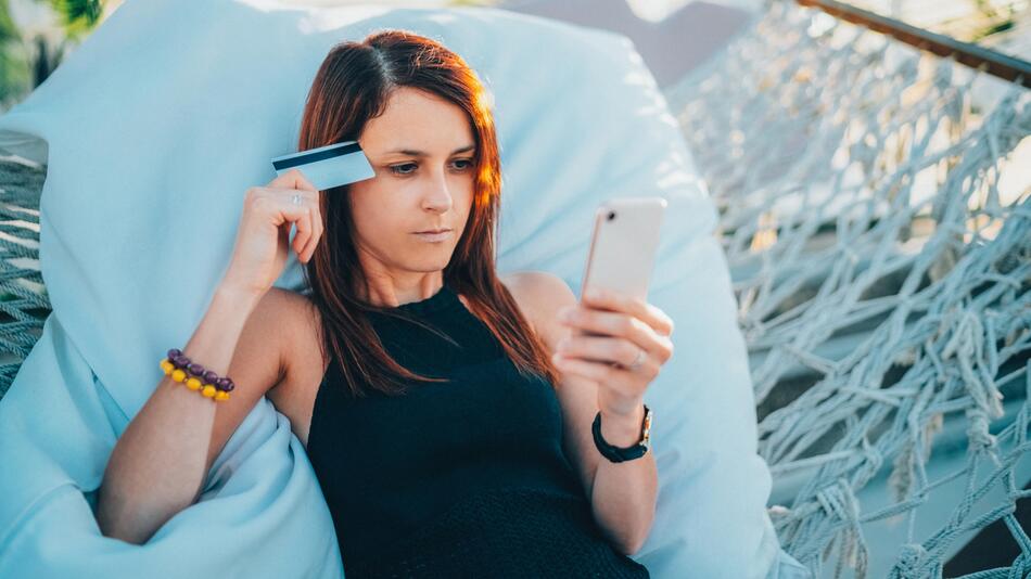 Frau schaut auf ihr Handy, hat eine kreditkarte in der Hand und liegt in einer Hängematte