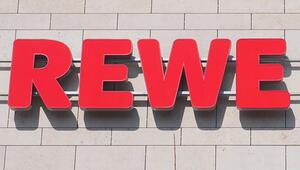Rote, große 3D-Buchstaben mit der Aufschrift "REWE" an einer hellen, gefliesten Außenwand.