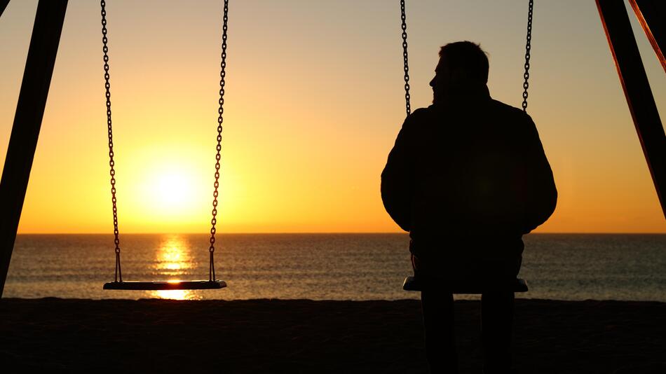 Mann sitzt auf Schaukel bei Sonnenuntergang neben leerer Schaukel