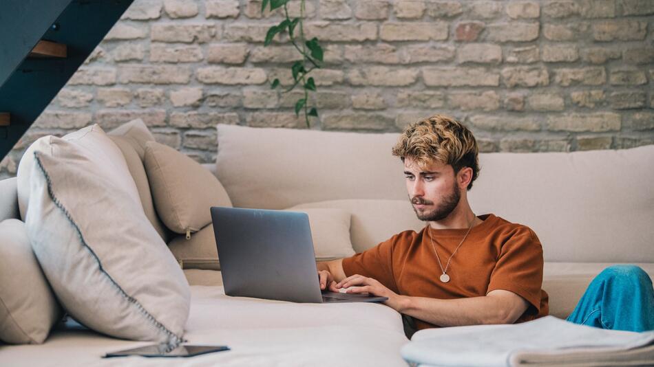 Ein Mann sitzt neben einer Couch und arbeitet am Laptop