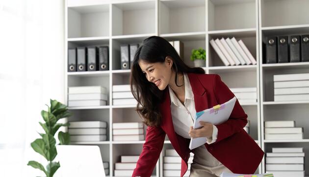 Eine lächelnde Frau hält zusammengeheftetes Papier in der Hand. Vor ihr liegt ein Papierstapel.