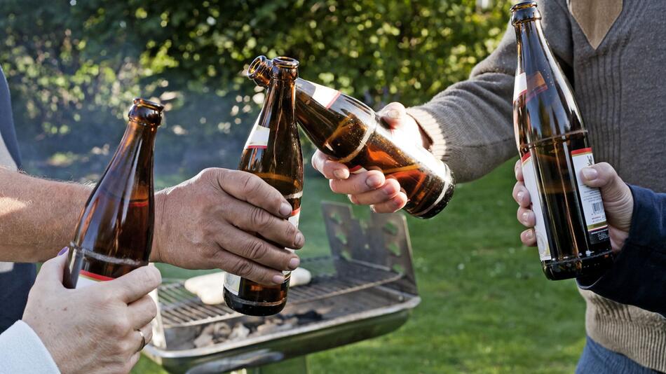 Vier Leute stoßen draußen vor einem Grill mit Bierflaschen an