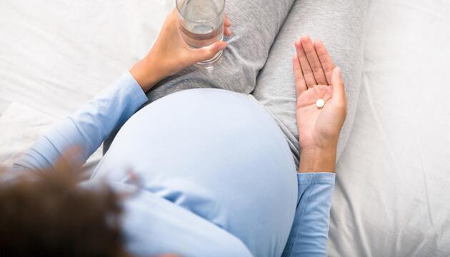 Schwangere Frau, die eine Tablette in der einen und ein Wasserglas in der anderen Hand hält