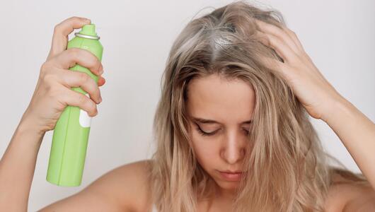 Junge blonde Frau mit schmutzigem, fettigem Haar, die ein Trockenshampoo auf die Haarwurzeln sprüht