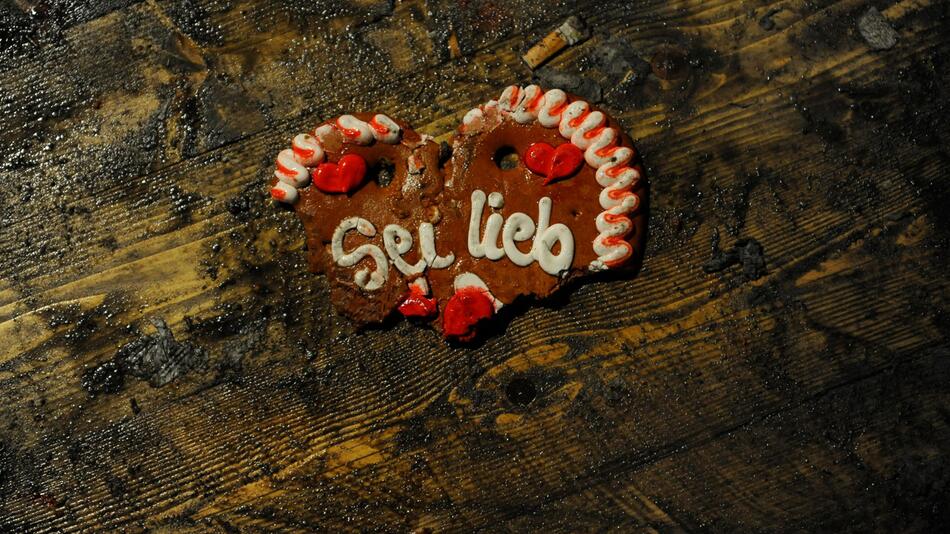 Ein zerbrochenes Lebkuchenherz auf einem Wiesnzeltboden. Die Schrift "Sie lieb" ist zu lesen.